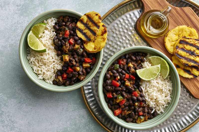Two mint-green ceramic bowls with white rice and black beans, garnished with lime and grilled plantains, arranged on a metal tray with a wooden cutting board and oil jar beside them