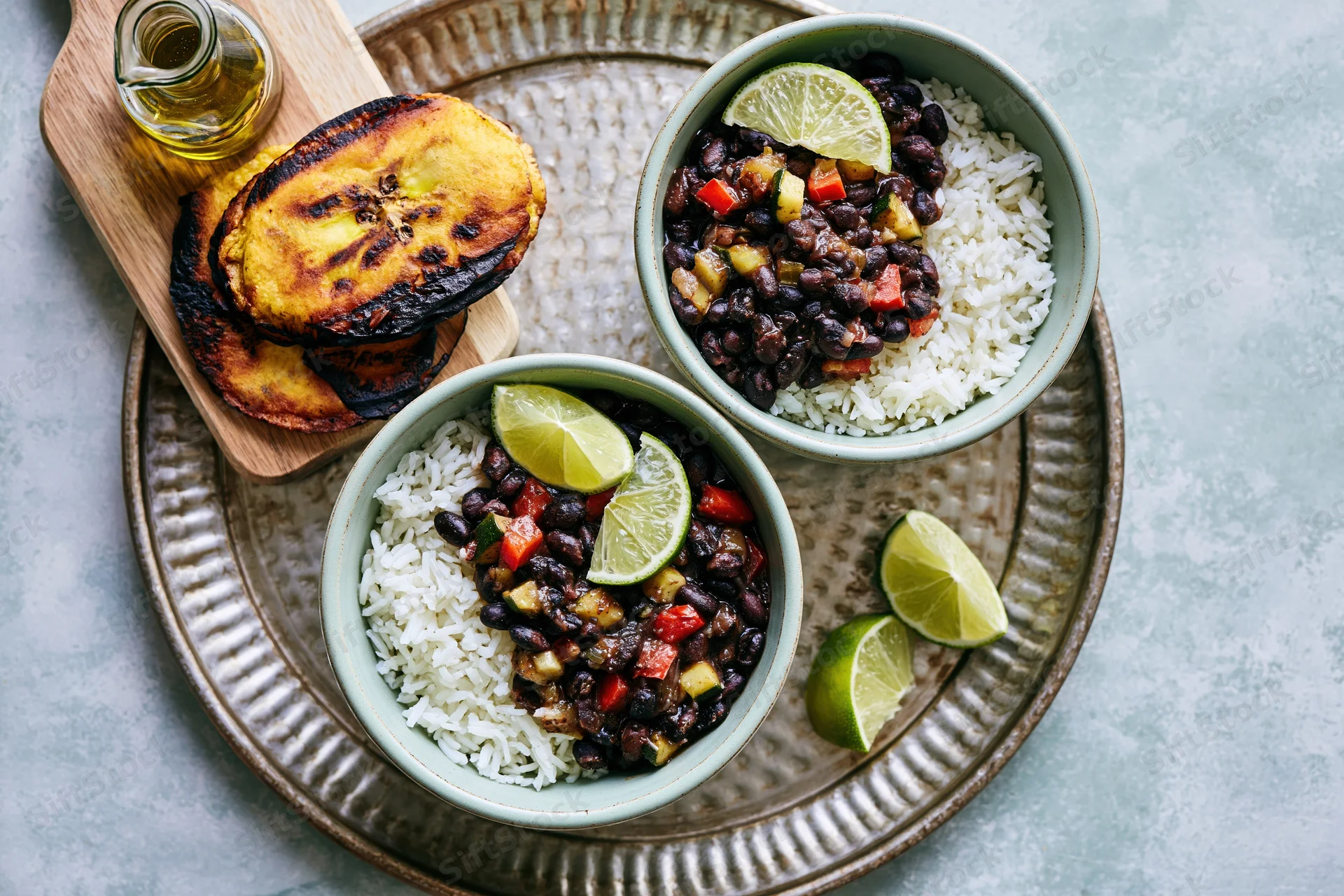 Two mint-green ceramic bowls with rice and black beans garnished with lime wedges on a textured tray with fried plantains and oil on the side