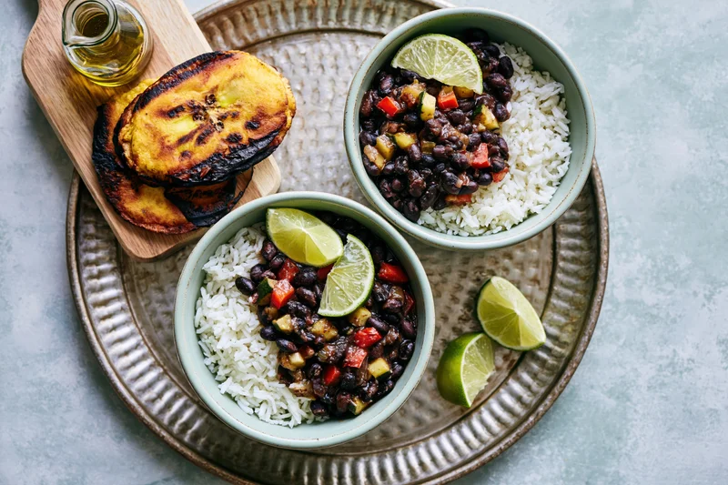 Two mint-green ceramic bowls with rice and black beans garnished with lime wedges on a textured tray with fried plantains and oil on the side