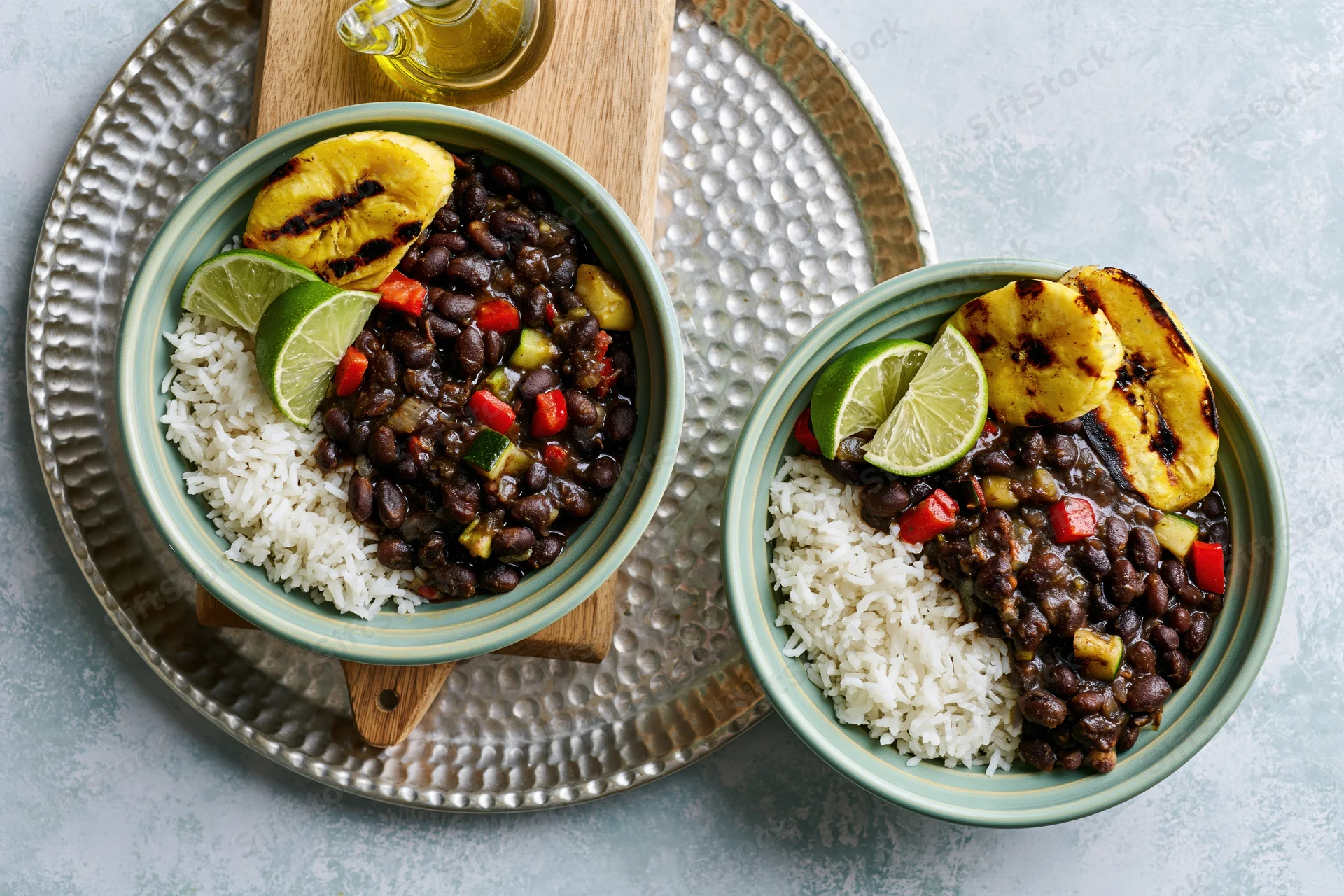 Two mint-green ceramic bowls with rice, black beans, grilled plantains and lime wedges on a hammered metal tray with a wooden board and oil glass nearby