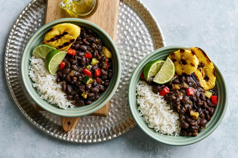 Two mint-green ceramic bowls with rice, black beans, grilled plantains and lime wedges on a hammered metal tray with a wooden board and oil glass nearby