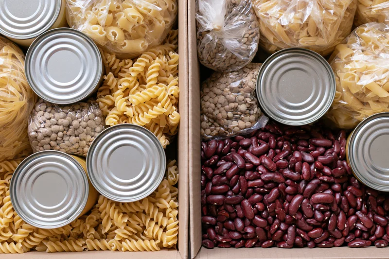 Two open cardboard boxes filled with assorted dry pantry goods including pasta, beans, and canned items