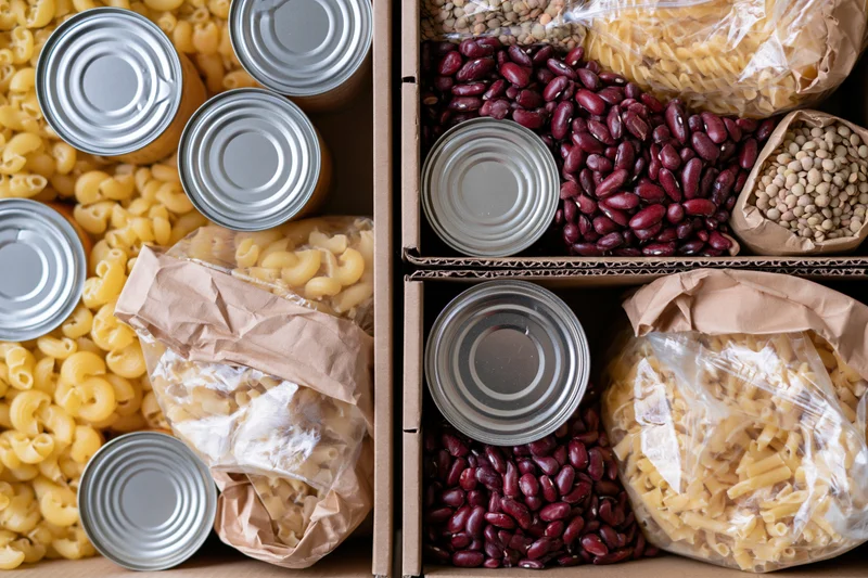 Open cardboard boxes filled with assorted dry pantry goods including canned beans, pasta, and legumes arranged for storage or donation
