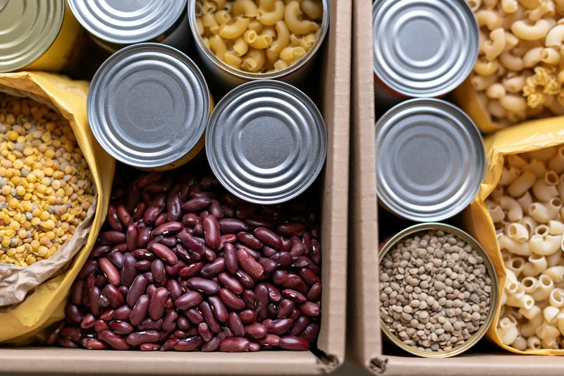 Two open cardboard boxes filled with assorted dry pantry goods including beans, pasta, corn, and lentils