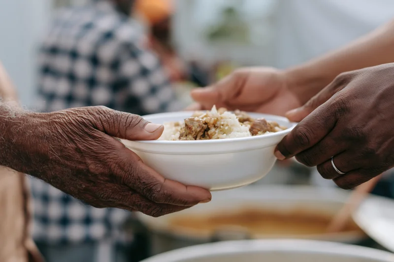 Weathered hands passing a white bowl of rice and meat between two people in a community kitchen