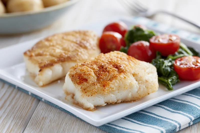 Two pan-seared white fish fillets with golden-brown crust served on a white plate with cherry tomatoes and spinach salad