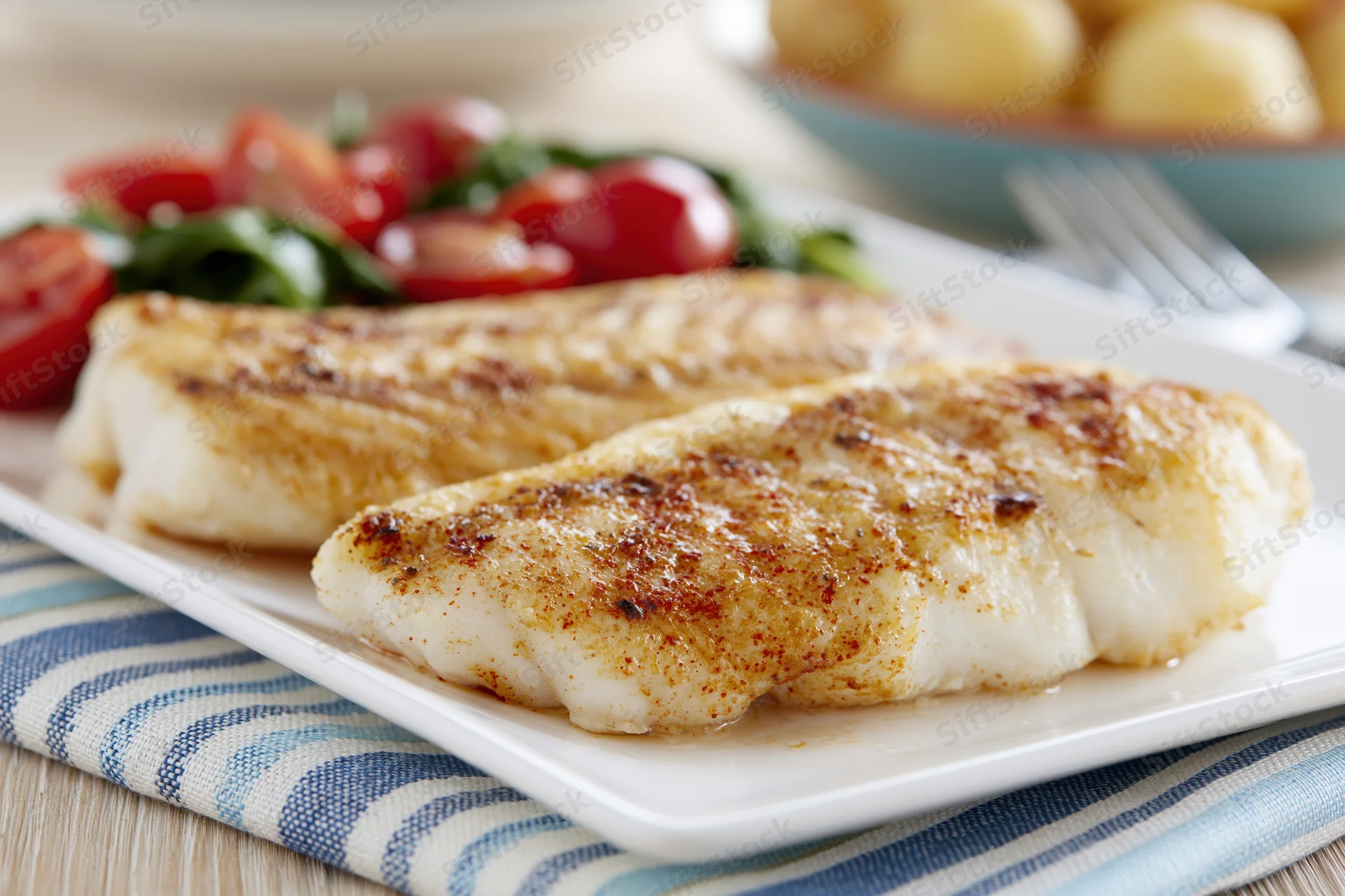 Two pan-seared white fish fillets with golden-brown crust served on a white plate with tomato salad and potatoes