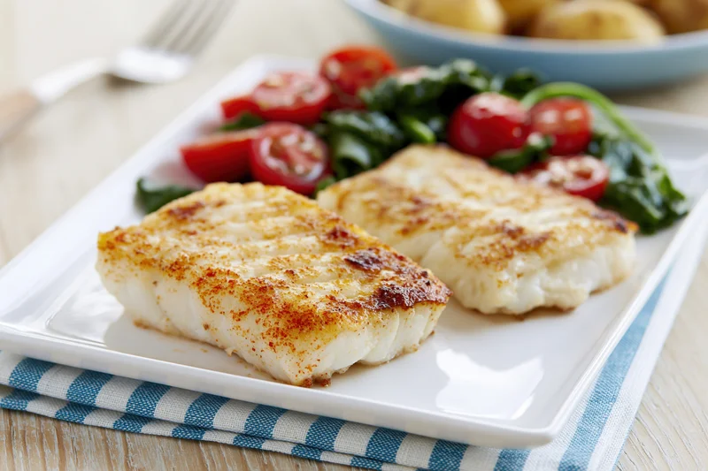 Two seared white fish fillets with tomato spinach salad on a white plate on a wooden table