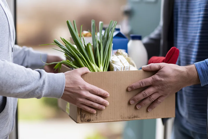 Two people exchanging a cardboard box of fresh groceries at a doorway
