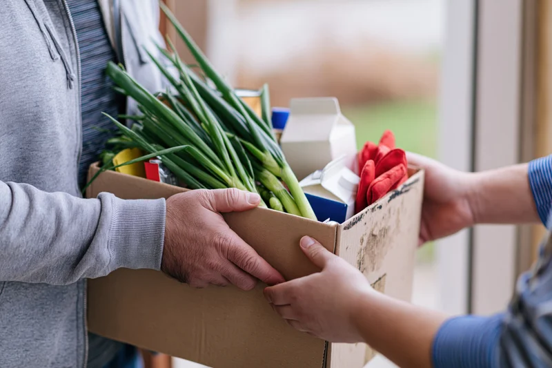 Two people exchanging a cardboard box of fresh vegetables and groceries at a doorway