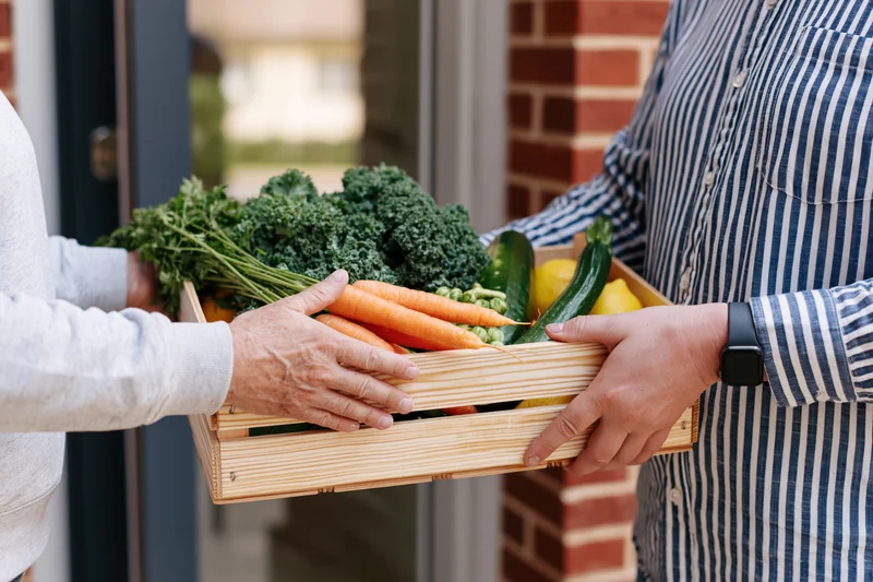 Two people exchanging a wooden crate of fresh vegetables at a front door