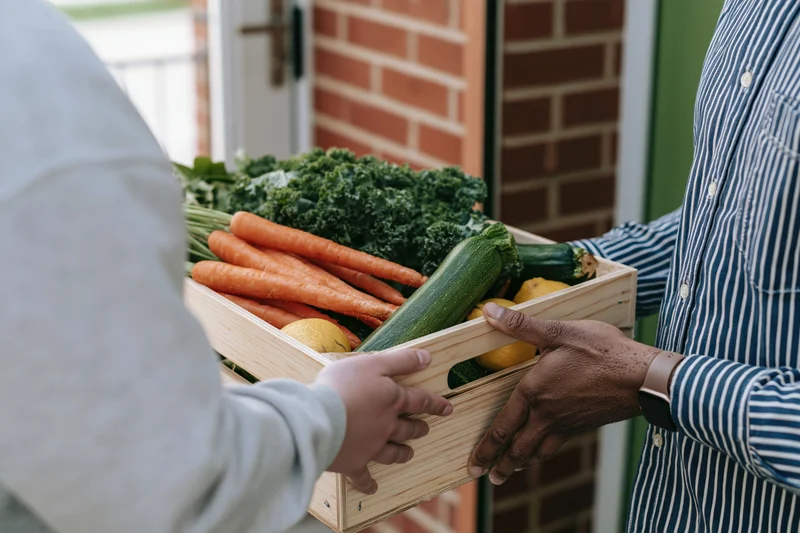 Two people exchanging a wooden crate of fresh vegetables at a front door