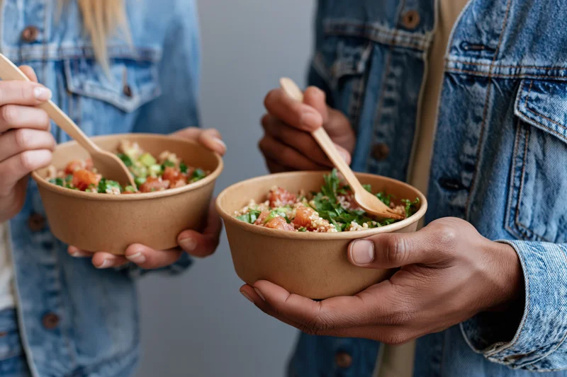 Two people holding kraft paper bowls of poke and grain salad with wooden spoons