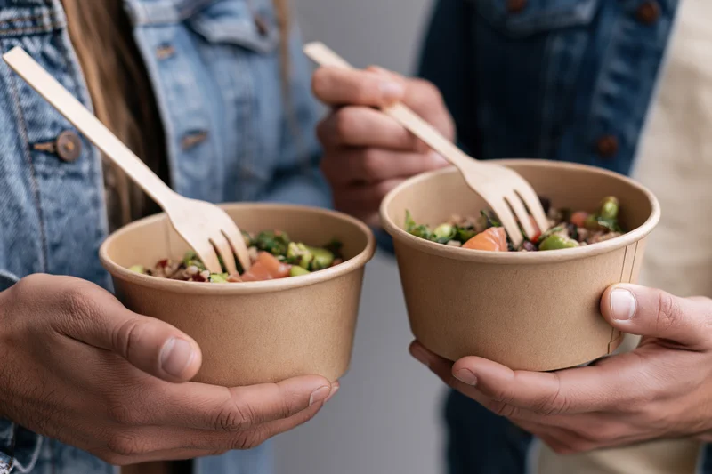 Two people holding kraft paper bowls of poke and grain salad with wooden forks, closeup view of hands and food