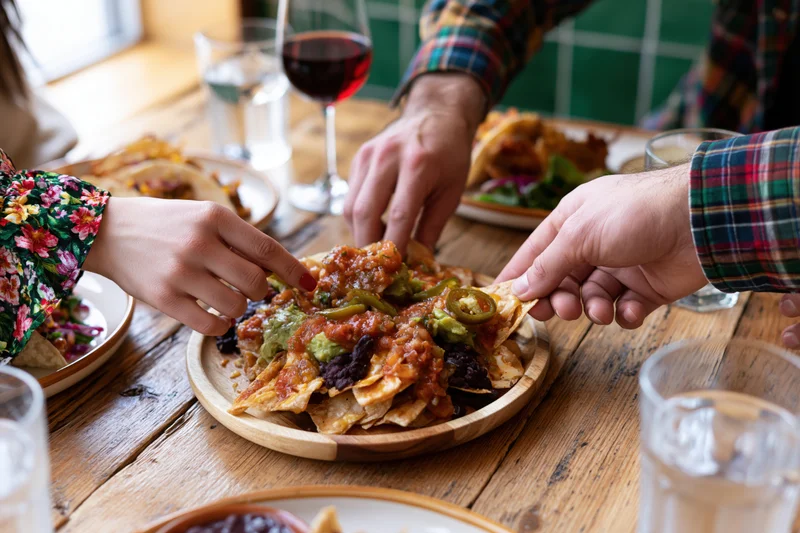 Two people reaching for loaded nachos on a wooden table in a restaurant
