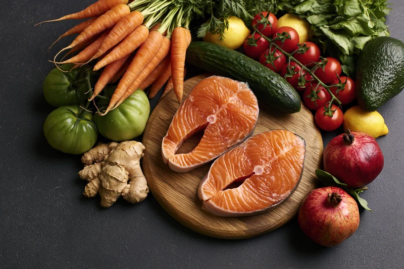 Two raw salmon steaks on a round wooden cutting board surrounded by fresh vegetables and fruits