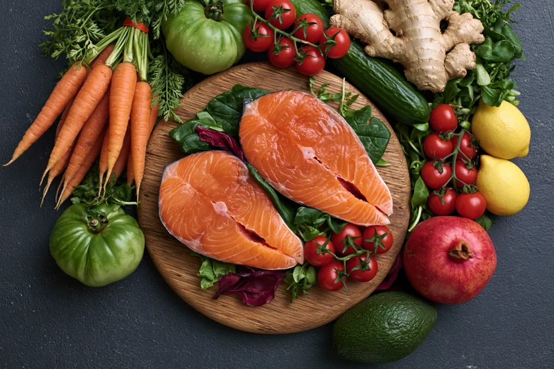 Two raw salmon steaks on a wooden cutting board surrounded by fresh vegetables and fruits