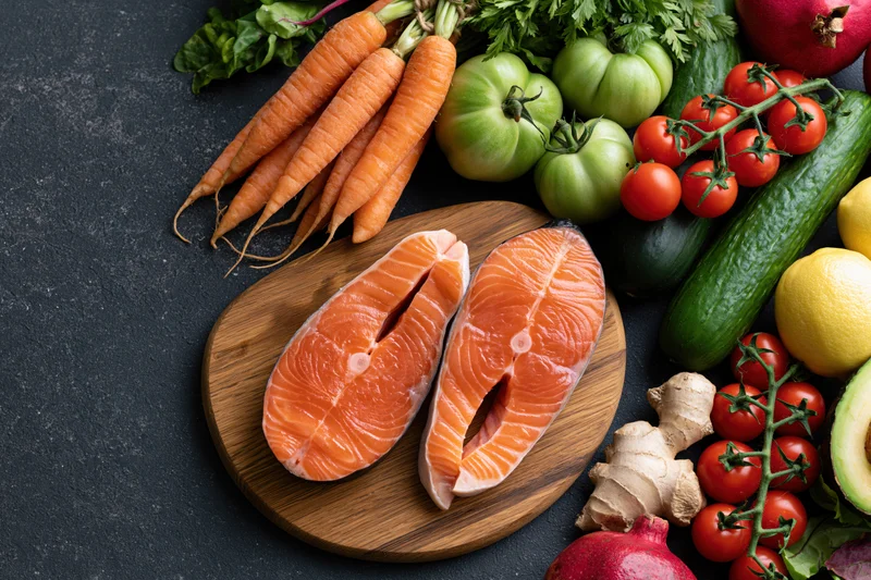 Two raw salmon steaks on a round wooden cutting board surrounded by fresh vegetables and herbs