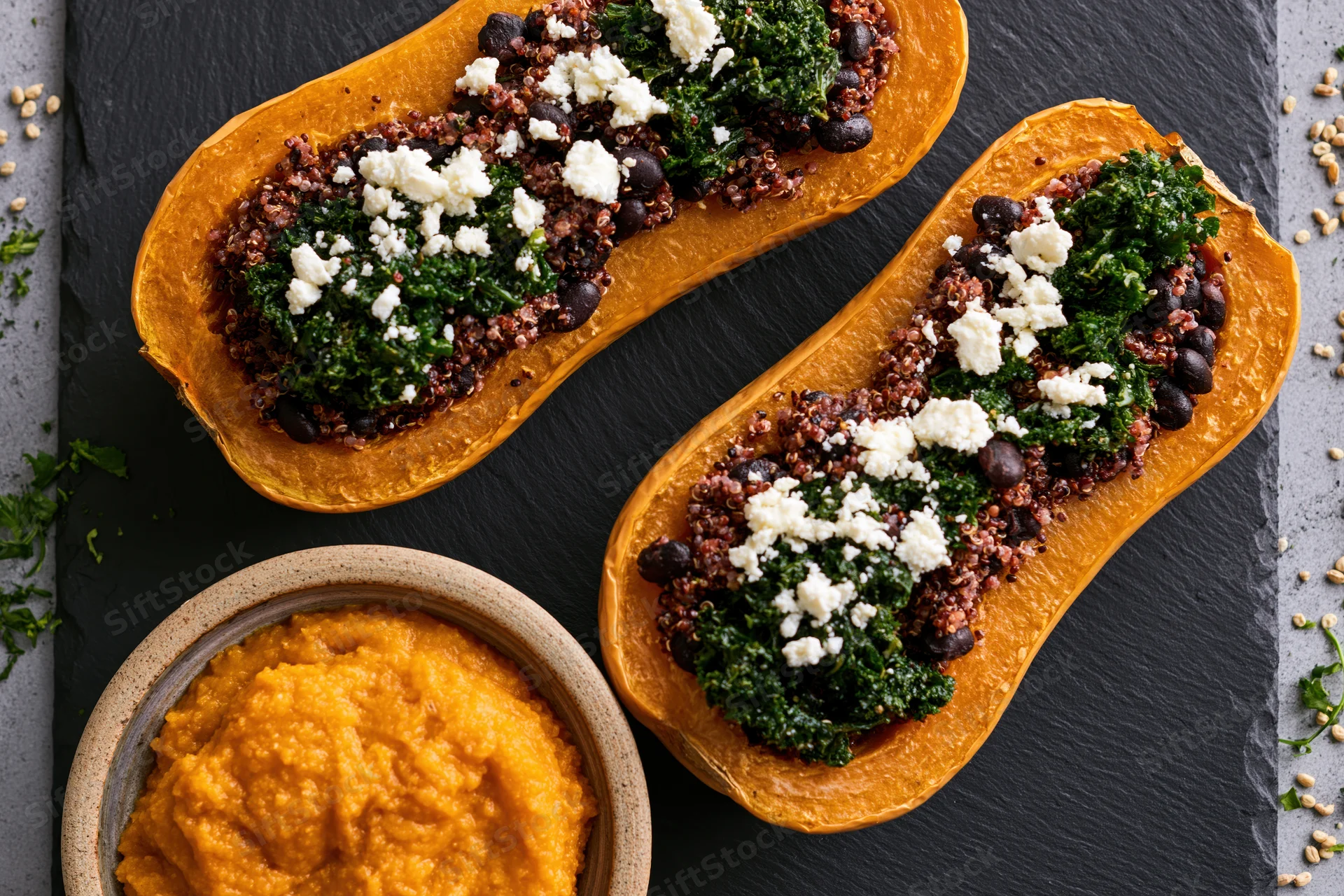 Two roasted butternut squash halves stuffed with quinoa kale and black beans on slate with side of squash puree in ceramic bowl