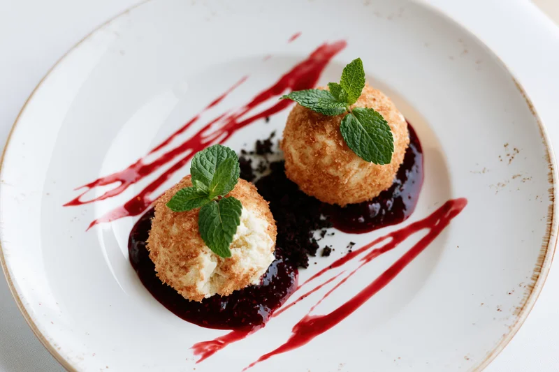 Two golden fried ice cream scoops with mint garnish on a white plate with red sauce drizzle