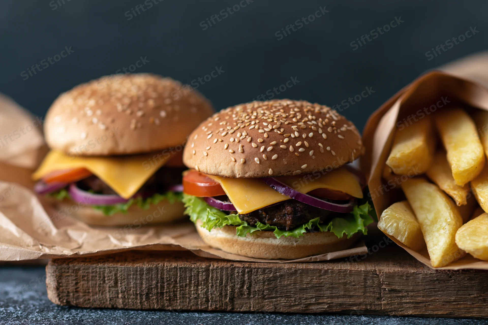 Two cheeseburgers with sesame seed buns and french fries on a wooden board