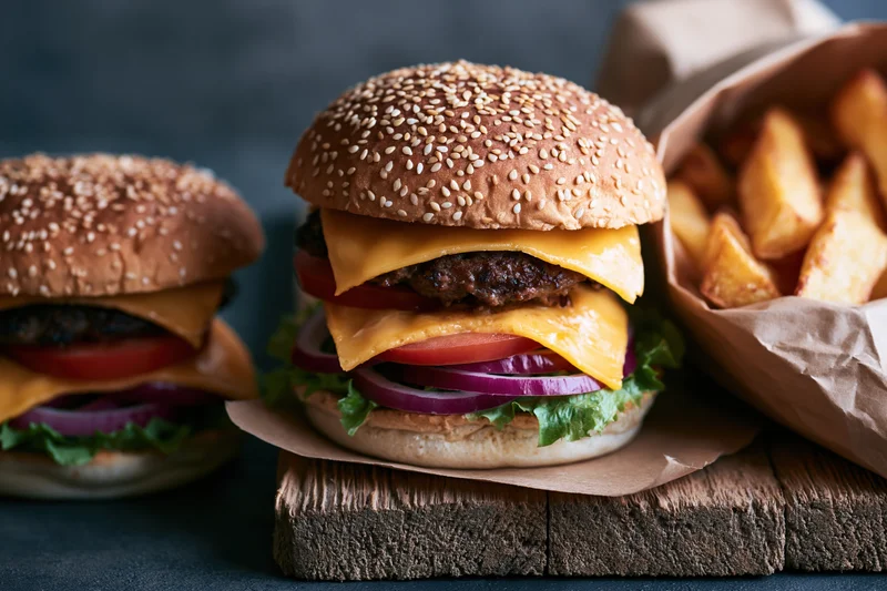 Two stacked cheeseburgers with sesame seed buns and french fries on a wooden board