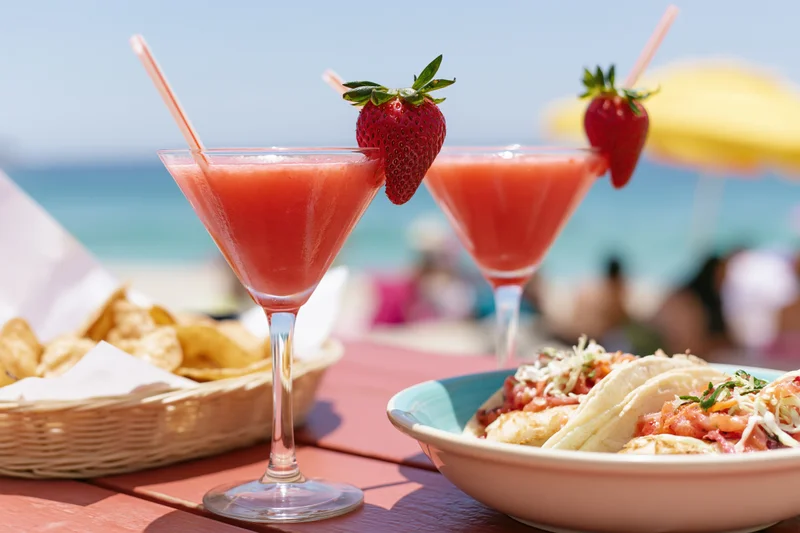 Two strawberry daiquiris in martini glasses with straws and strawberries on a beach table beside tacos and chips