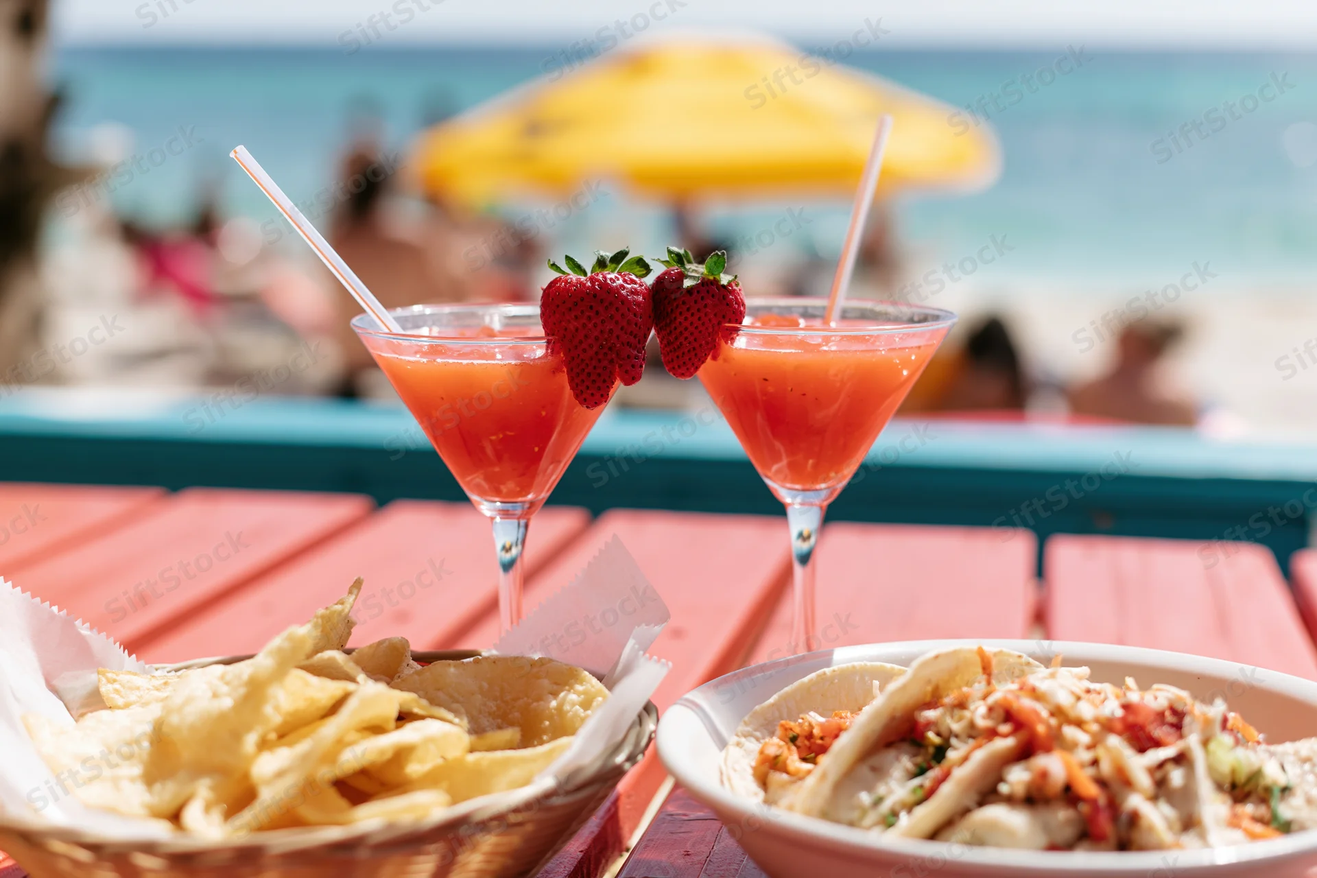 Two strawberry daiquiris in martini glasses with straws and fruit garnish on a wooden table beside tacos and chips at a beachside bar