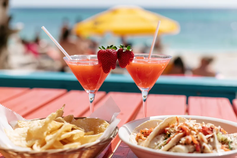 Two strawberry daiquiris in martini glasses with straws and fruit garnish on a wooden table beside tacos and chips at a beachside bar
