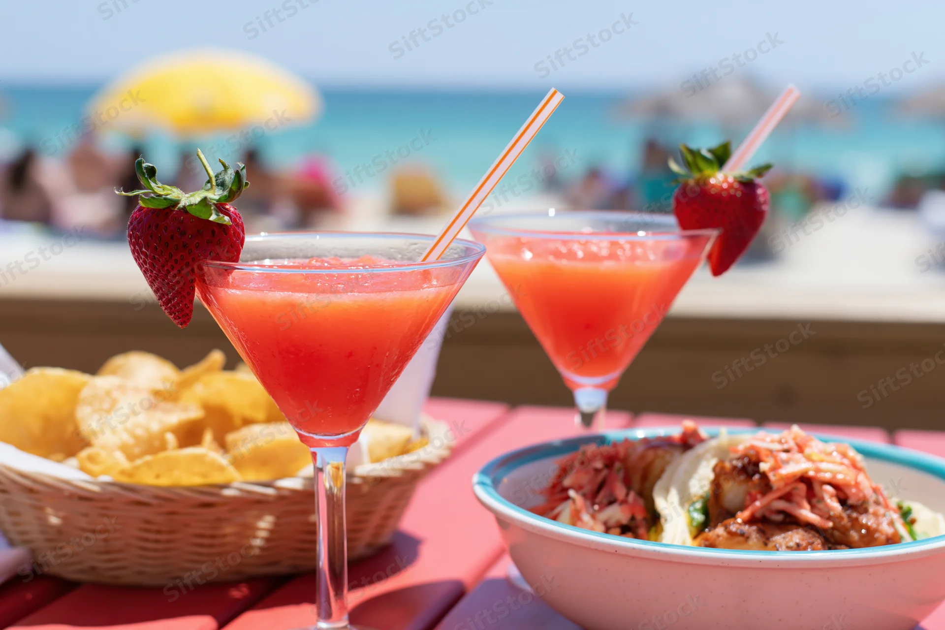 Two strawberry daiquiris in martini glasses with straws and garnishes on a beach table beside chips and tacos