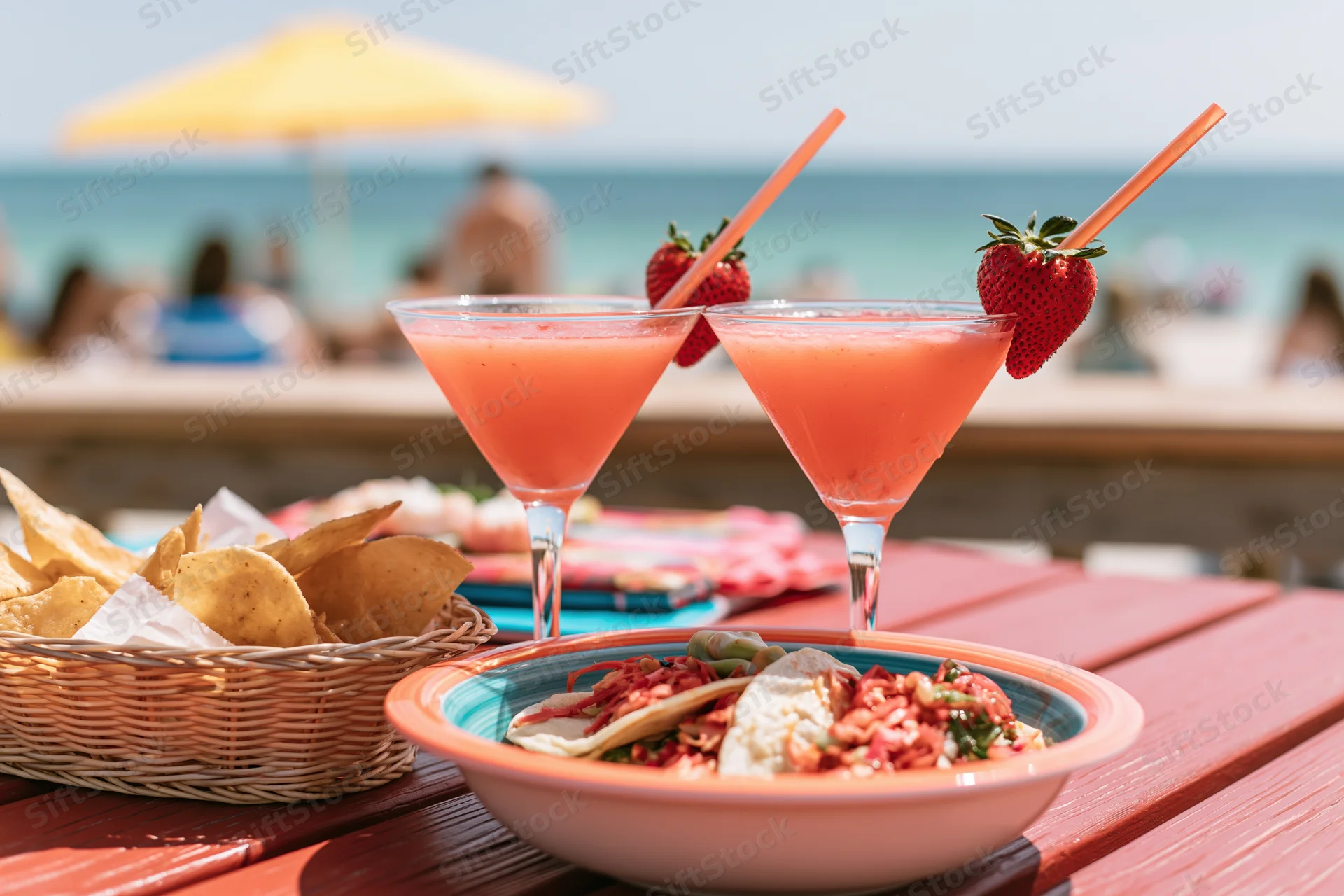 Two strawberry daiquiris in martini glasses with straws and fruit garnishes on a beach table beside a bowl of tacos and a basket of chips