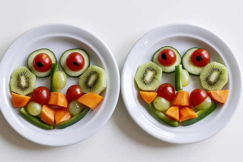 Two white plates with fruits and vegetables arranged into smiling faces on a white surface