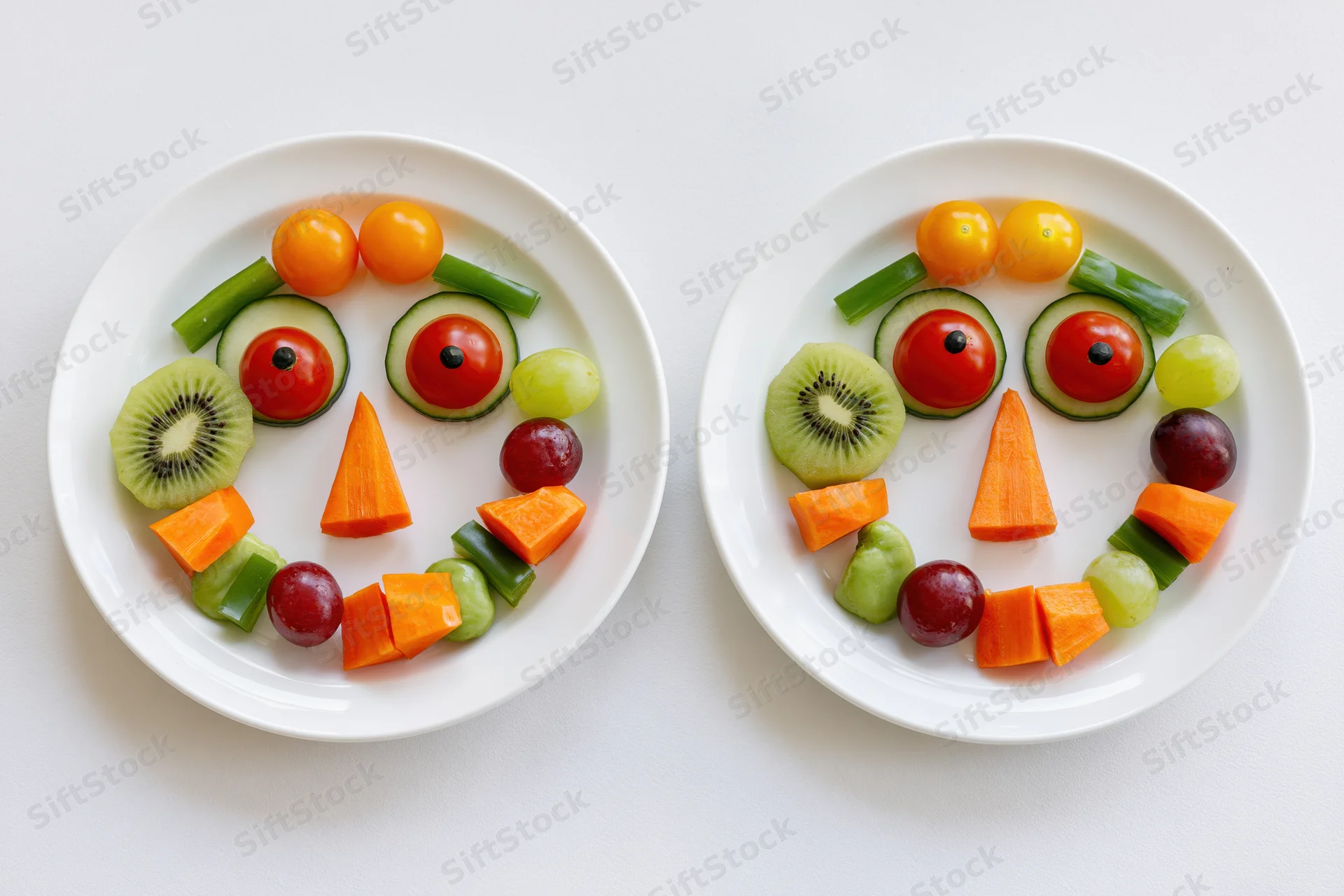 Two white plates with fruits and vegetables arranged into smiling faces side by side on white surface