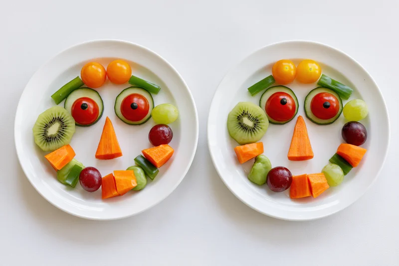 Two white plates with fruits and vegetables arranged into smiling faces side by side on white surface