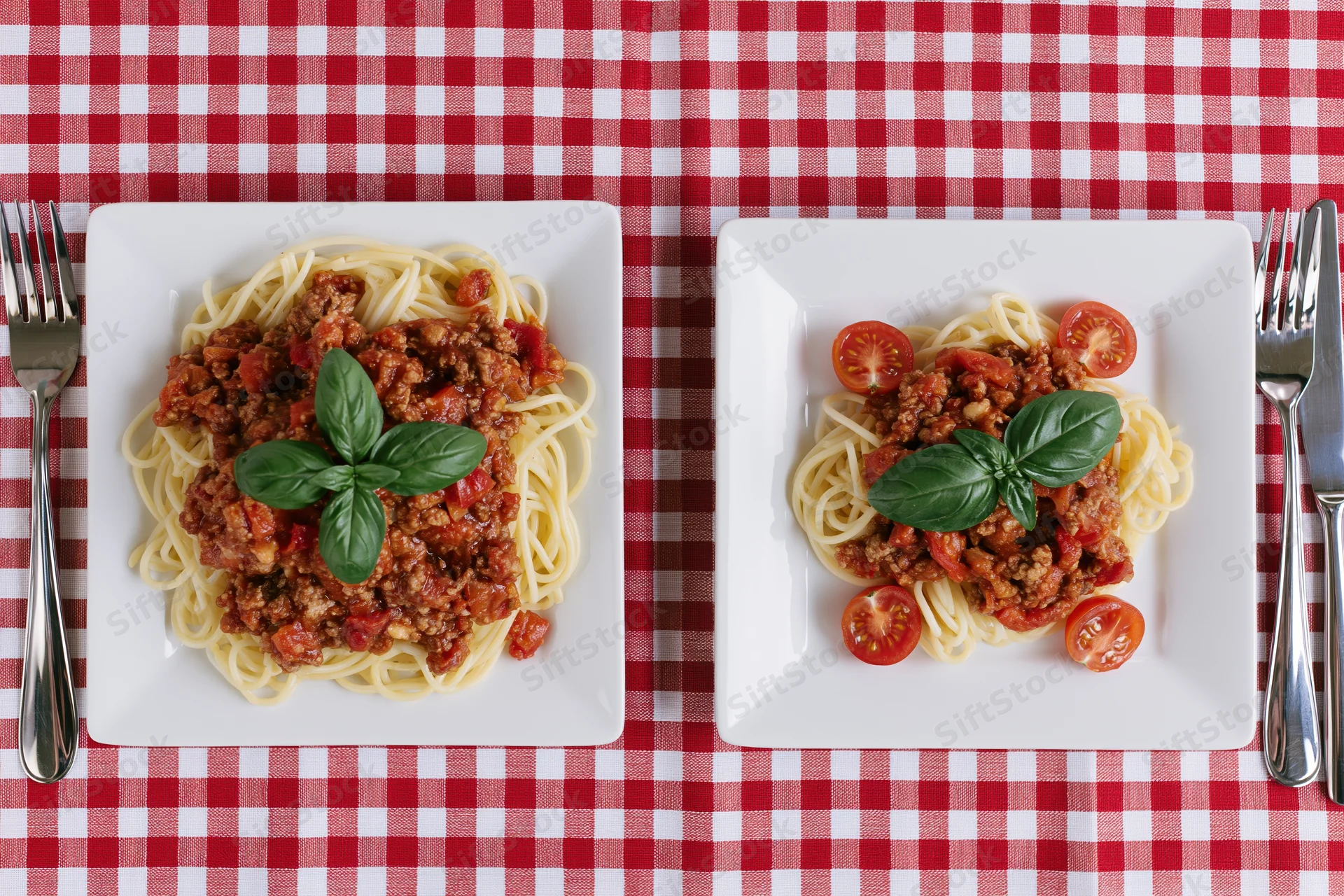 Two square white plates with spaghetti bolognese and basil on a red and white checkered tablecloth, one larger portion beside a smaller one, viewed from above with forks and knives on either side