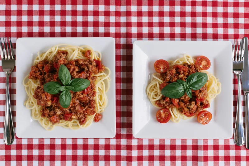 Two square white plates with spaghetti bolognese and basil on a red and white checkered tablecloth, one larger portion beside a smaller one, viewed from above with forks and knives on either side