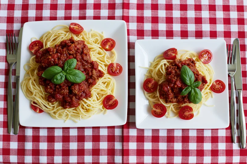 Two square white plates with spaghetti bolognese and basil on a red and white checkered tablecloth