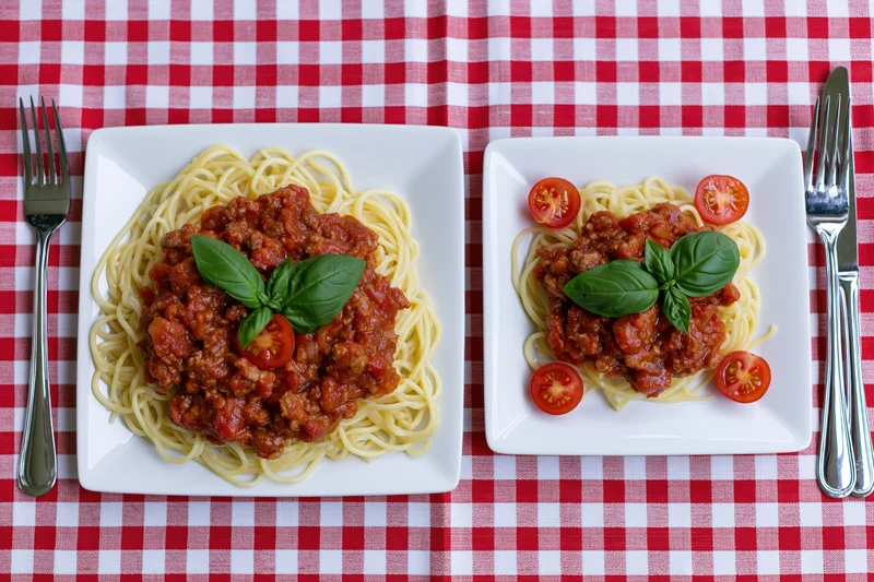 Two square white plates with spaghetti bolognese and basil on a red and white checkered tablecloth