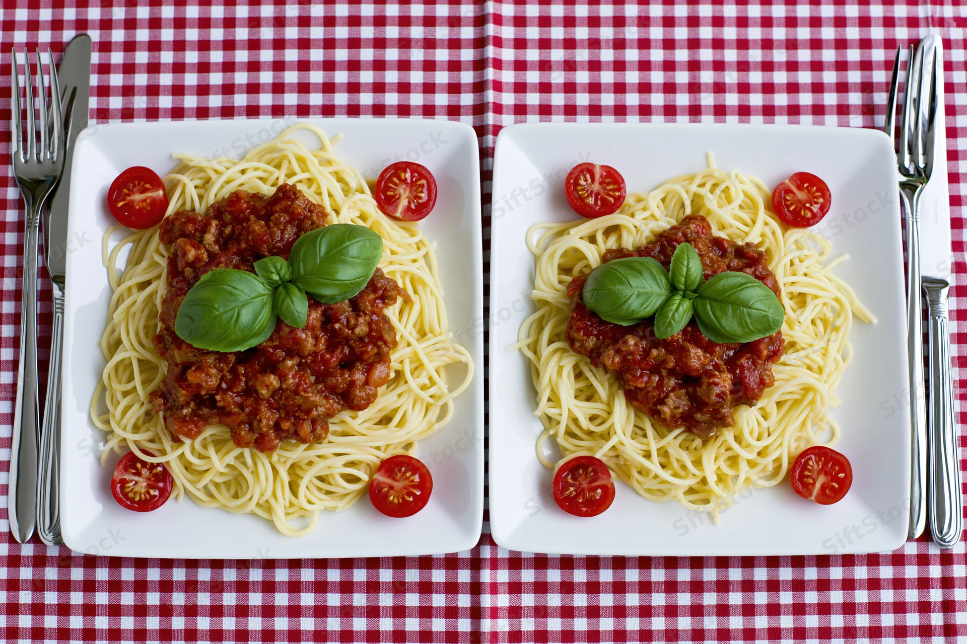 Two square white plates with spaghetti bolognese garnished with basil and tomato halves on a red and white checkered tablecloth
