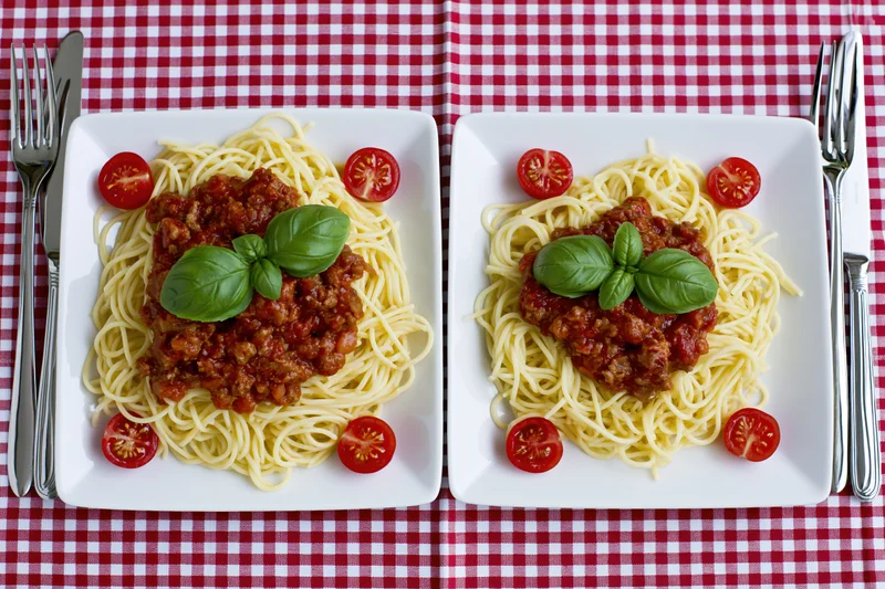 Two square white plates with spaghetti bolognese garnished with basil and tomato halves on a red and white checkered tablecloth