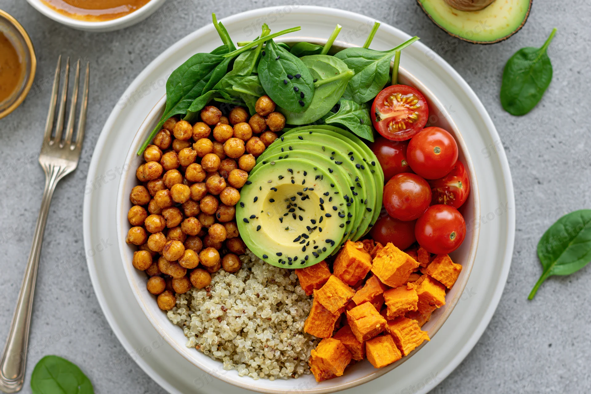 Vegan buddha bowl with quinoa, roasted chickpeas, avocado, cherry tomatoes, spinach and sweet potato on a white plate viewed from above