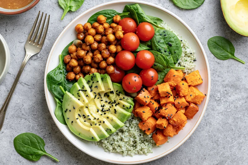 Vegan buddha bowl with avocado, quinoa, chickpeas, tomatoes, spinach and sweet potato on white ceramic plate viewed from above on gray surface