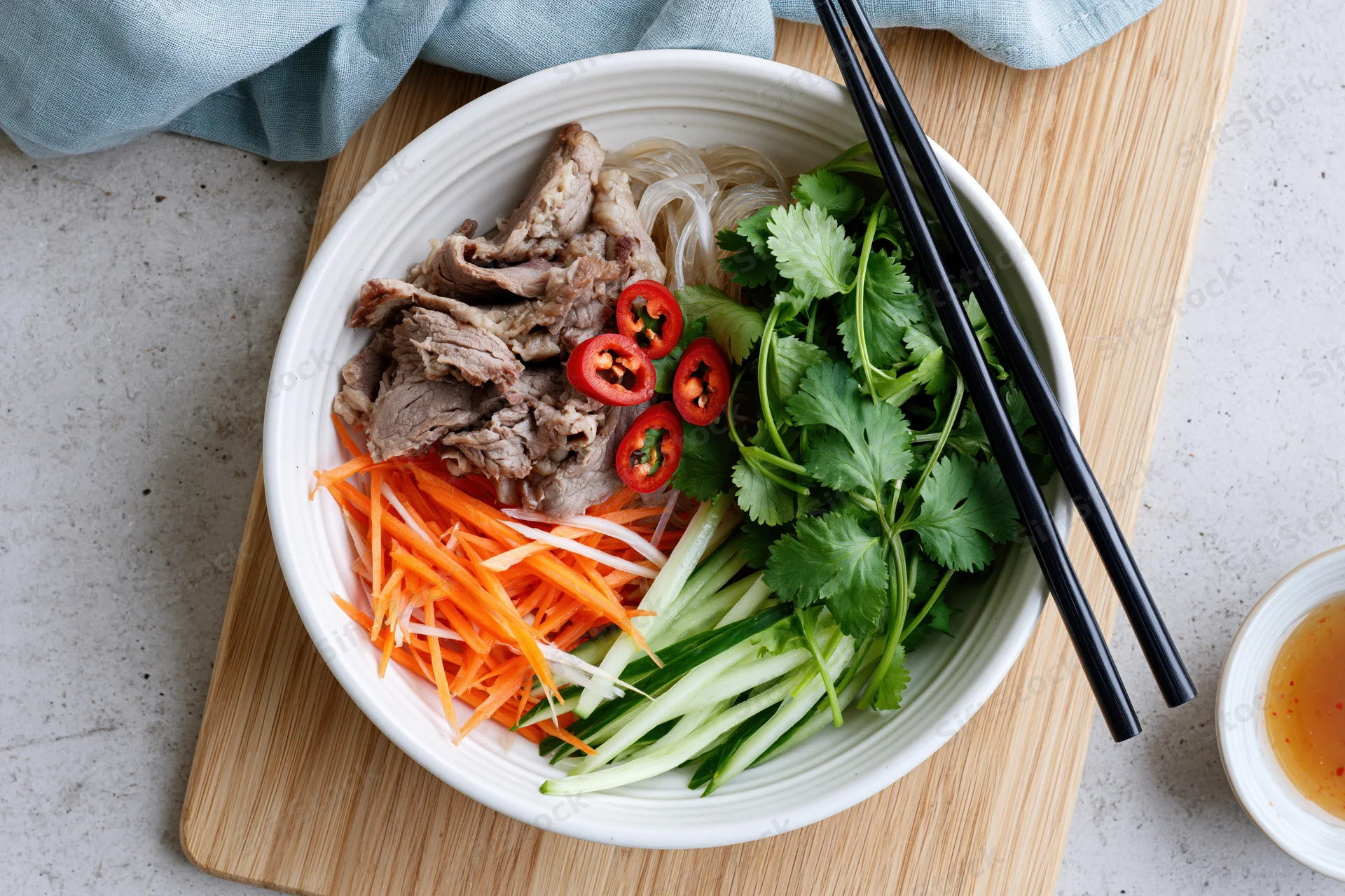 Vietnamese noodle bowl with sliced beef, shredded carrot, cilantro, cucumber and chili on wooden cutting board