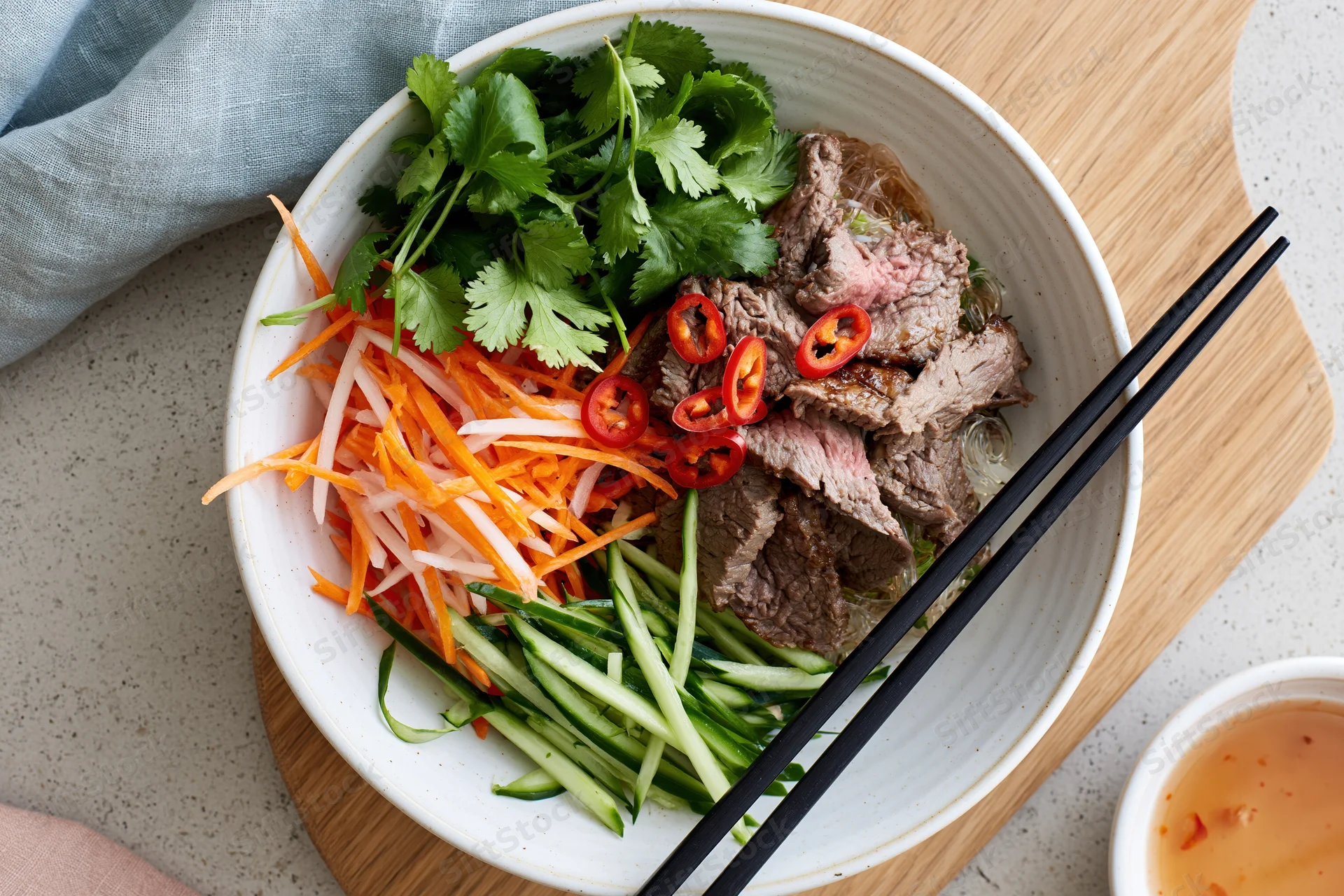 Vietnamese noodle bowl with sliced beef, shredded carrots, cilantro, cucumber, and chili, served with chopsticks on a wooden board