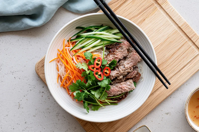 Vietnamese noodle bowl with sliced beef, shredded carrot, cilantro, and chili on a bamboo cutting board