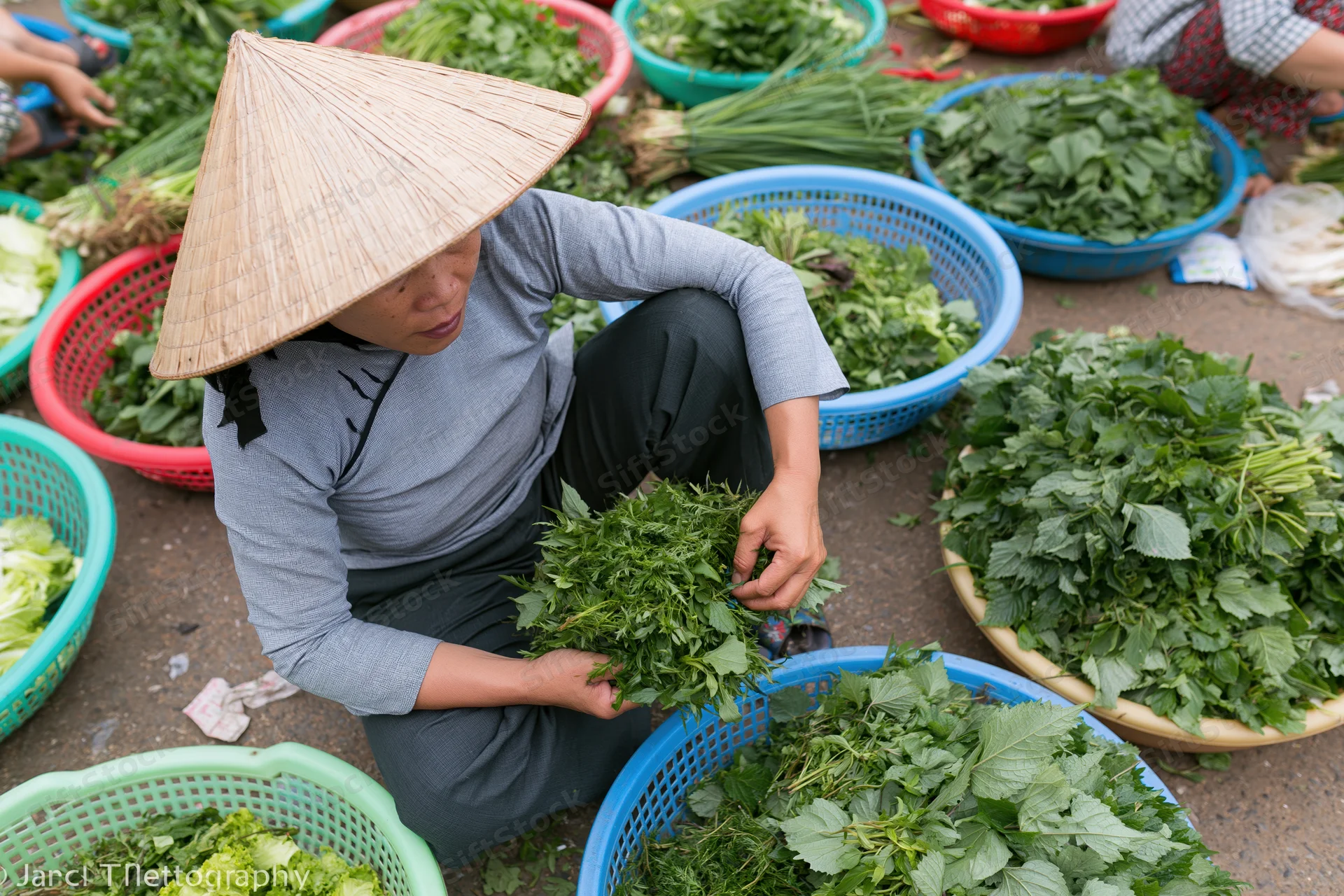 Vietnamese woman sorting fresh leafy vegetables at outdoor market while crouching among colorful baskets