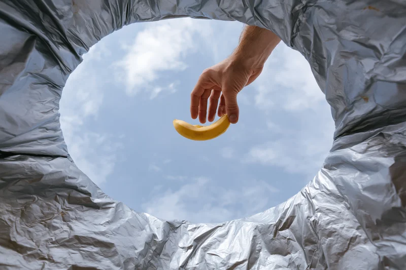 Hand dropping a banana peel into a trash bin viewed from inside looking up