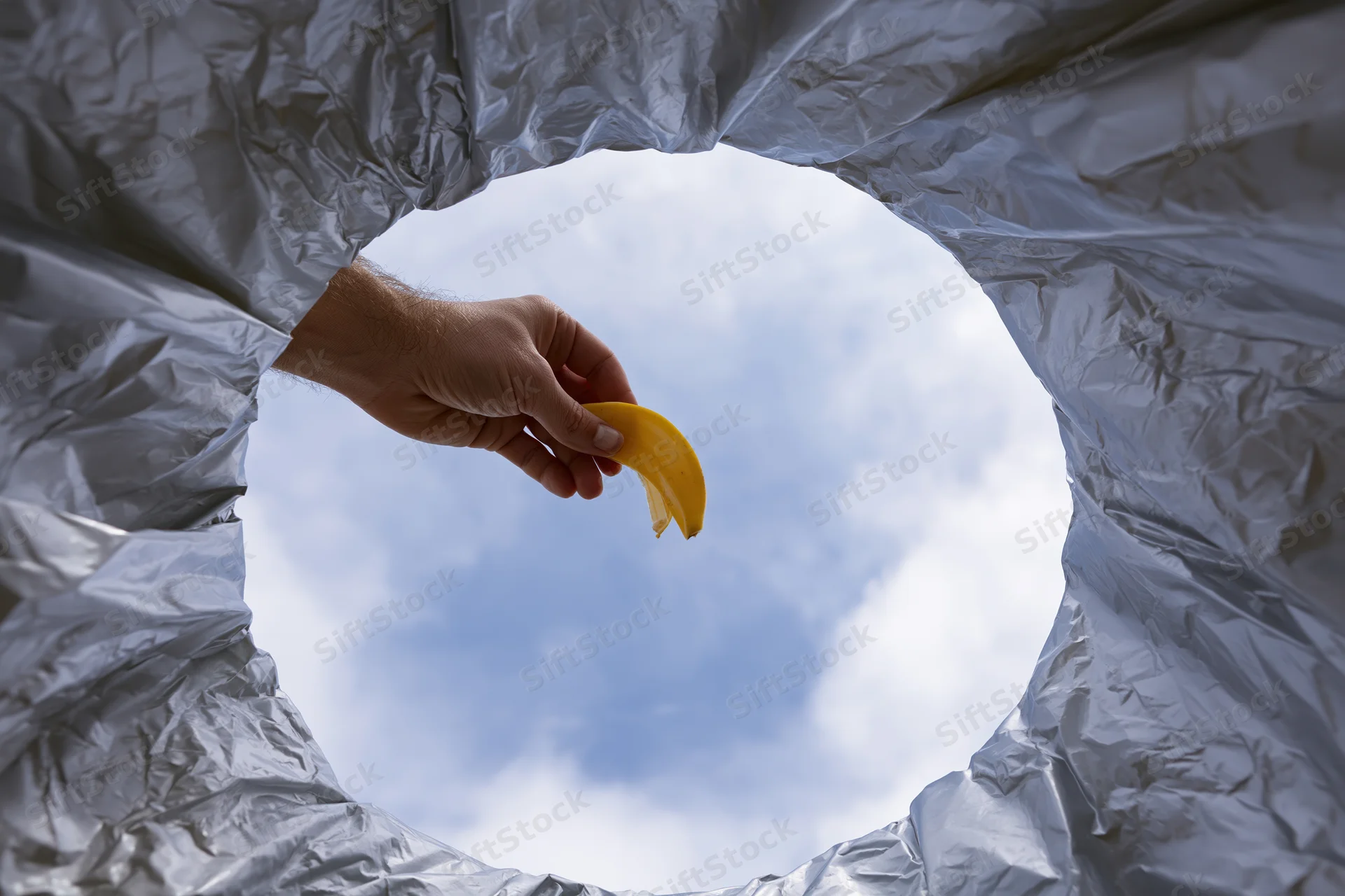 Hand dropping banana peel into trash bin viewed from inside looking up at sky