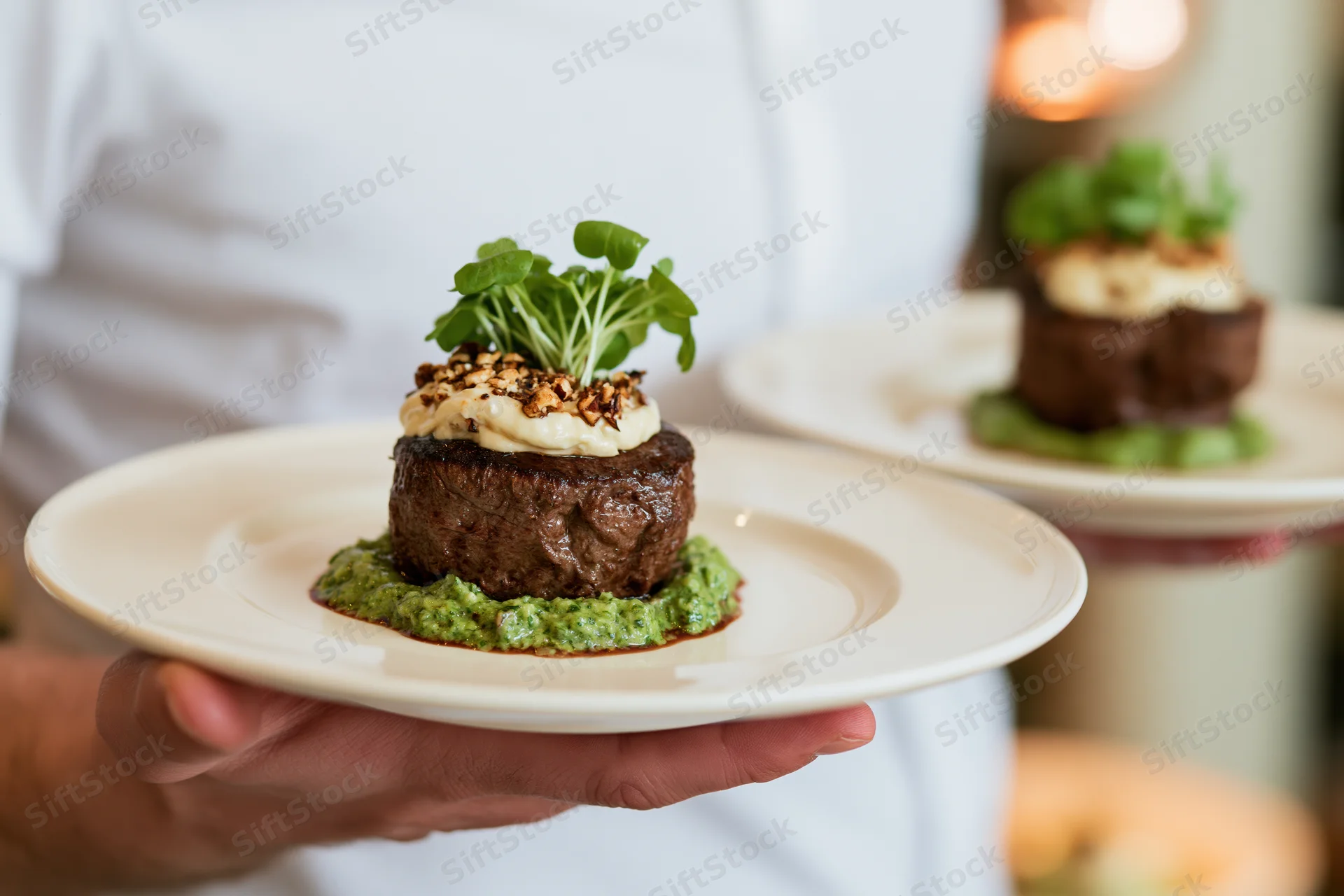 Waiter holding two plates with seared steak dishes garnished with cream and microgreens