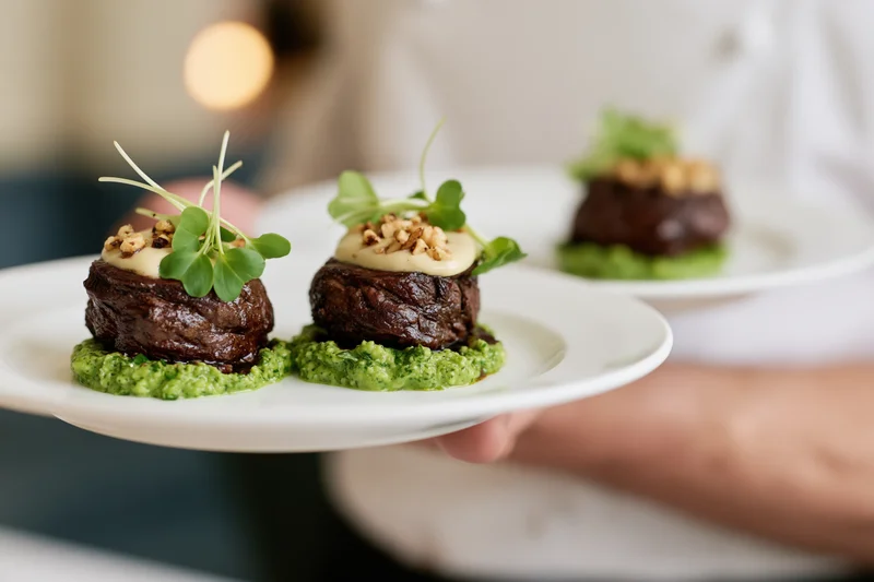 Waiter holding two plates with seared beef appetizers on green sauce garnished with microgreens and nuts